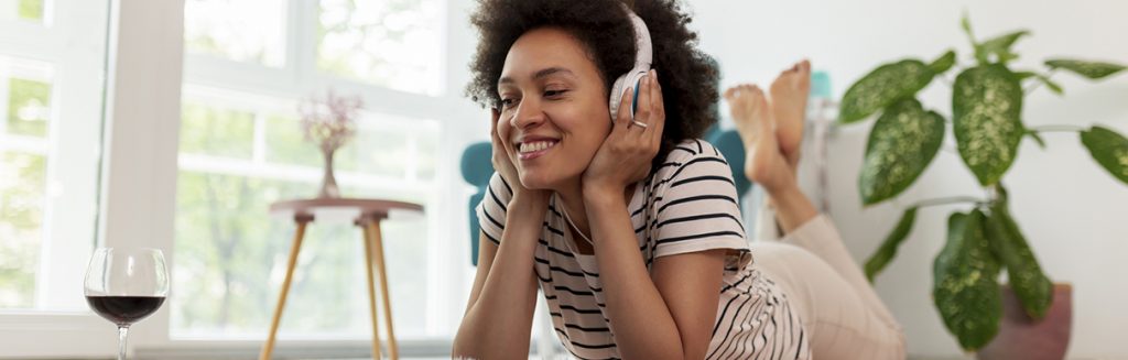 Mulher feliz com fone de ouvido deitada no chão de casa e uma taça de vinho ao lado
