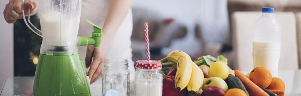 Cozinha com leite vegetal no liquidificador e em garrafas e frutas diversas na fruteira