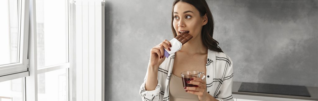 Mulher comendo um chocolate e com uma xícara de café na outra mão