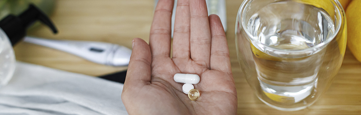 Mão segurando diversos comprimidos com um copo de água ao lado