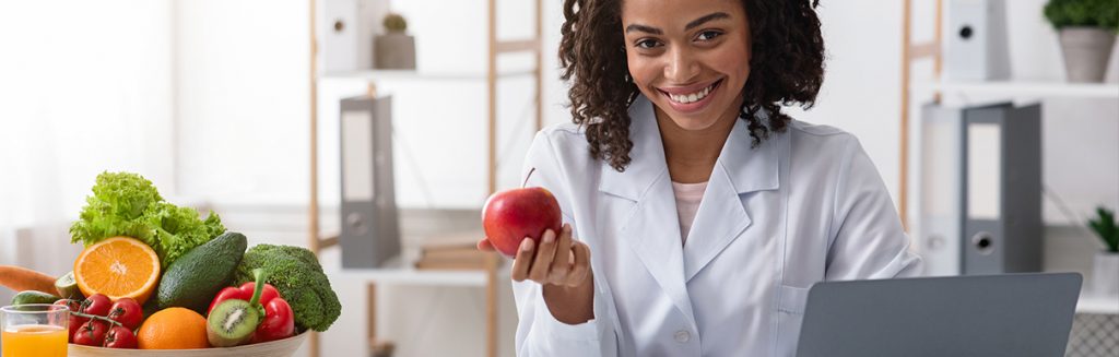 Nutricionista sorrindo com maçã na mão e outros alimentos saudáveis no consultório 