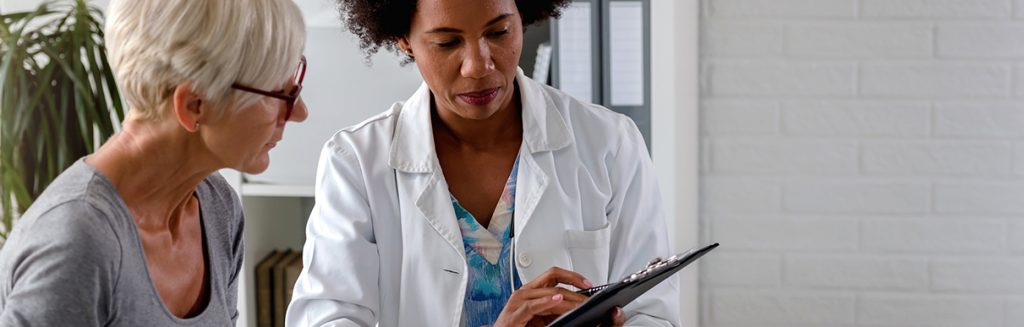 Senhora olhando para prancheta que mulher de jaleco branco mostra a ela: consulta médica