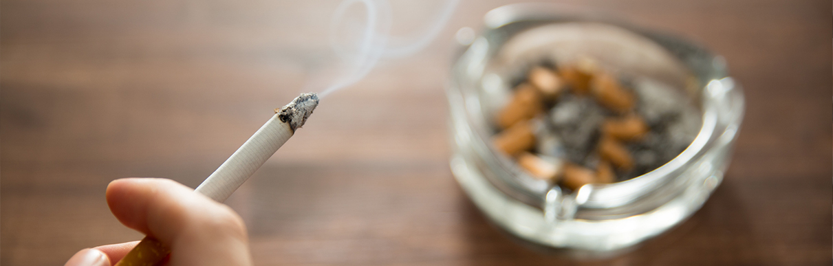 Cigarro aceso na mão de uma pessoa com cinzeiro ao fundo