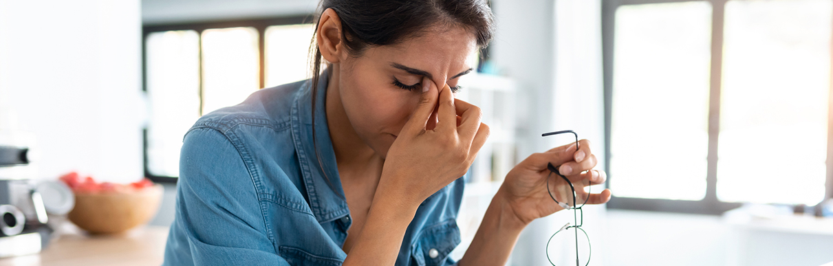 Mulher com os óculos em uma mão e segurando o rosto em outra, simbolizando o estresse