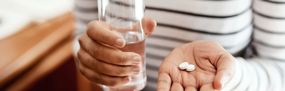 Pessoa segurando dois comprimidos em uma mão e um copo de água em outra